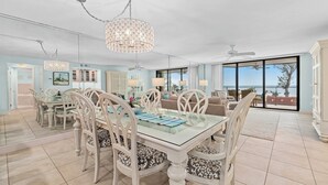 A spacious dining area with a glass table and white chairs, a chandelier, and a view of the ocean through large windows.