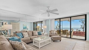 A spacious living room with a large window offering a view of the beach, a ceiling fan, and a comfortable seating area.
