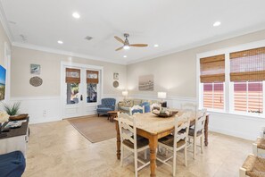 Un coin repas avec une table et des chaises en bois, un ventilateur de plafond et une vue sur l’extérieur à travers une fenêtre munie de stores.