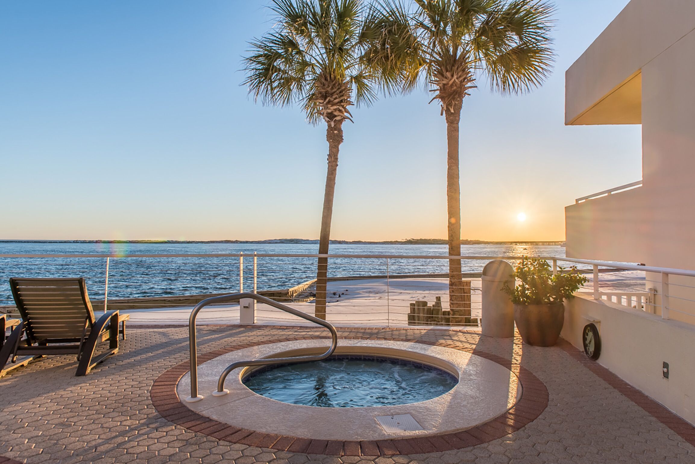 A rooftop pool area with a hot tub, lounge chairs, and palm trees overlooking the sea.
