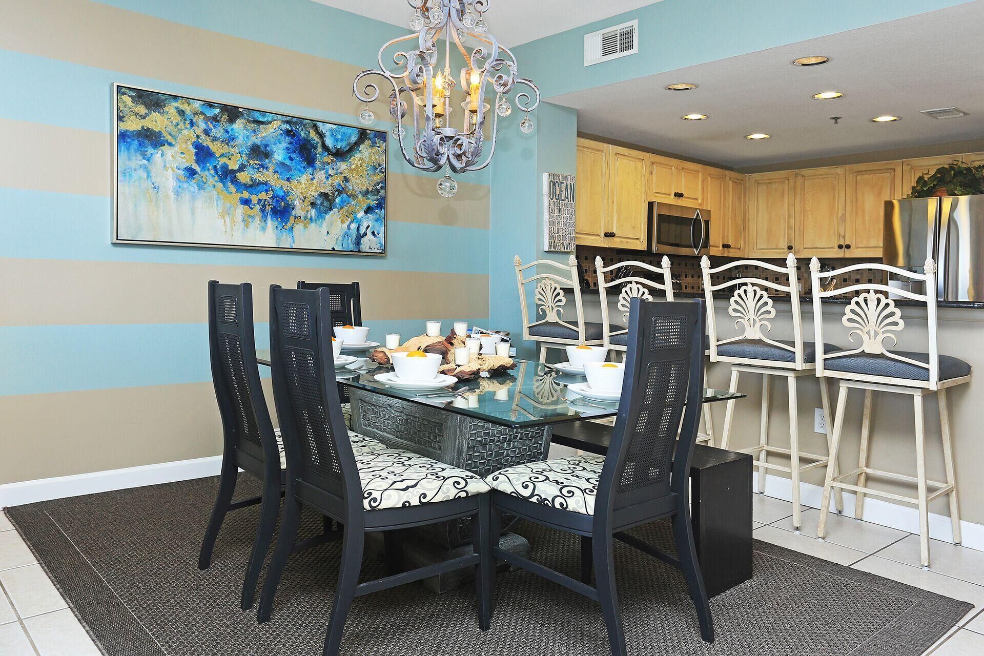 A dining area with a glass table, black chairs, and a chandelier. There is a painting on the wall and a kitchen in the background.