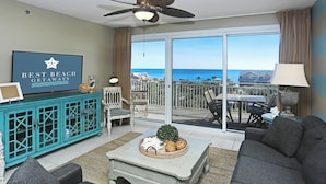 A living room with a view of the ocean, a ceiling fan, a teal cabinet, and a television displaying 'Best Beach Getaways'. - GULF VIEWS IN A  PREMIER RESORT!! GREAT RATES! (Destin)