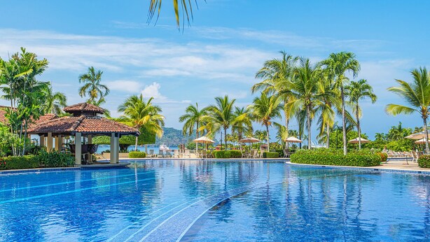 Una piscina amplia y transparente con sillas de descanso y sombrillas, rodeada de palmeras y un pabellón.