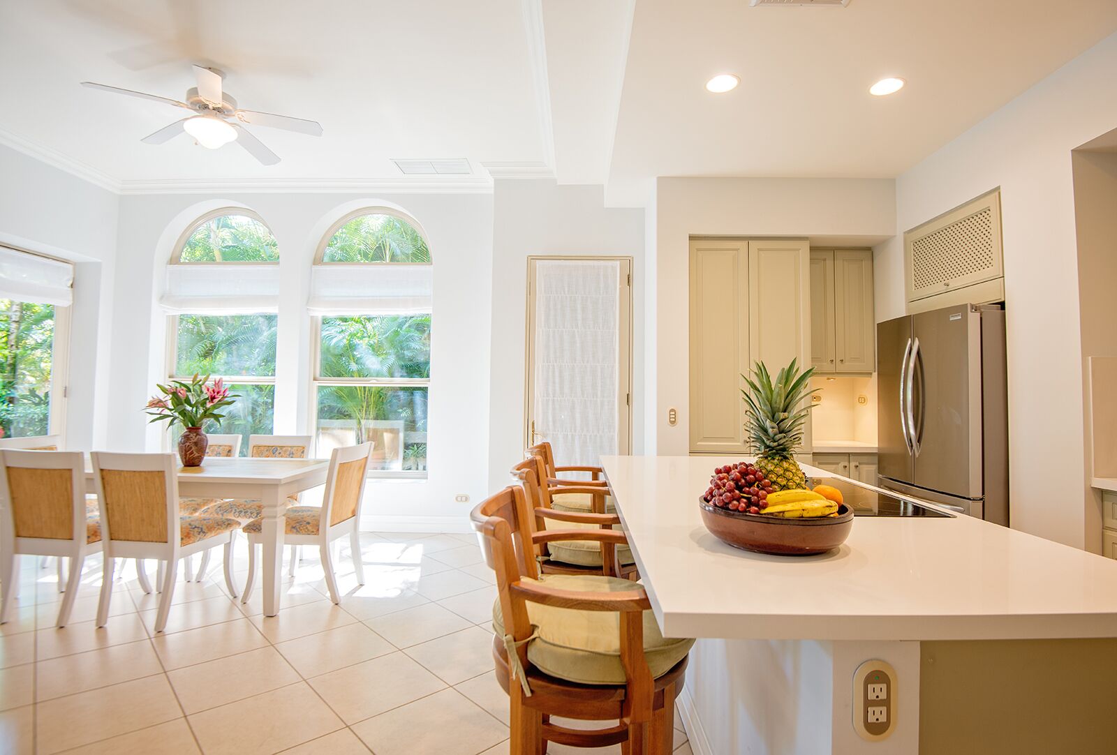 A bright kitchen with a central island, a dining area with chairs, and large windows with a view of greenery.
