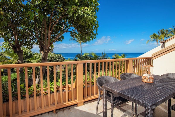A balcony with a wicker table and chairs, overlooking a view of the ocean and palm trees.