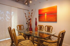 A dining area with a glass table surrounded by four chairs, a painting of a sunset, and decorative vases.