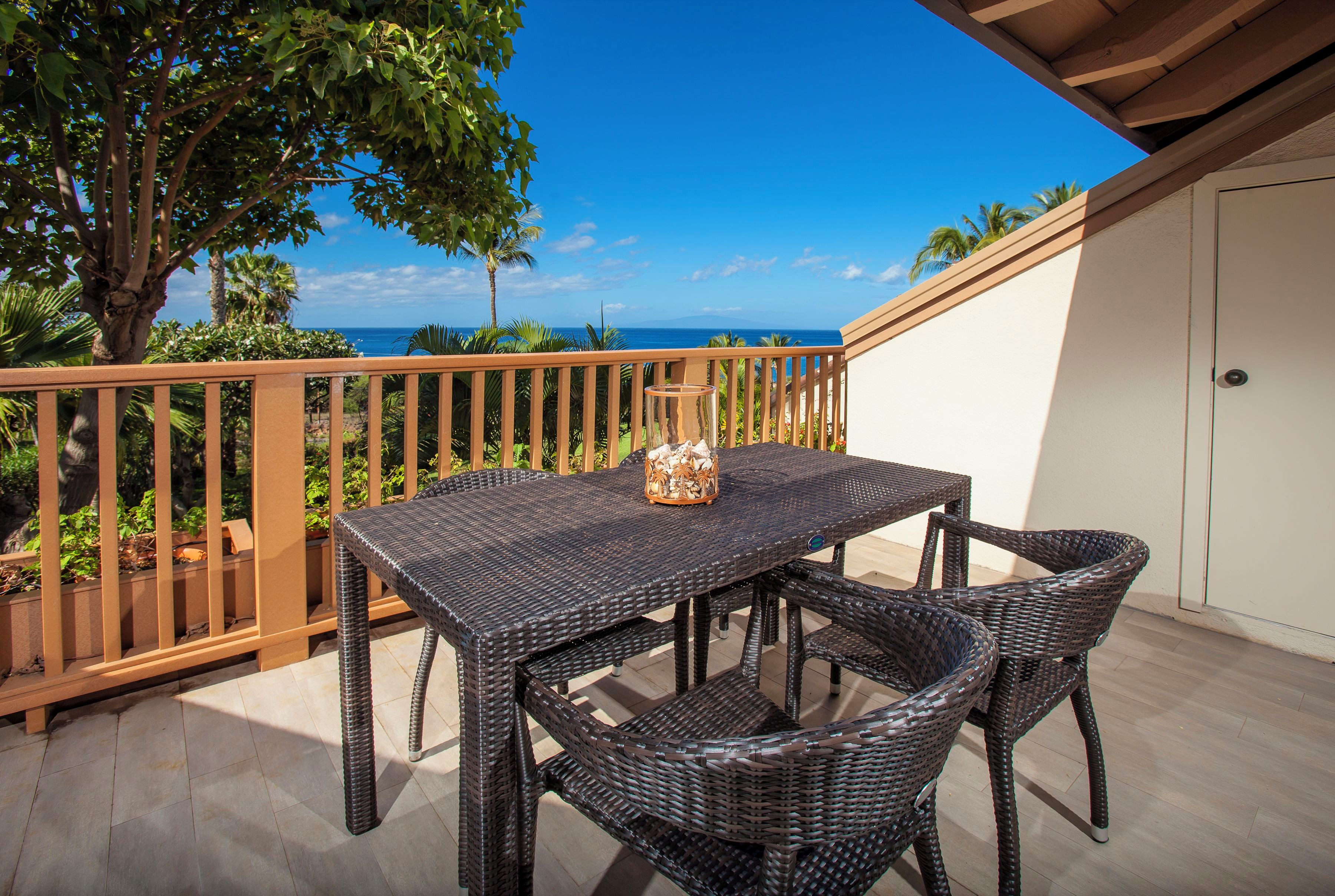 A balcony with a wicker table and chairs, overlooking a view of the ocean and palm trees.