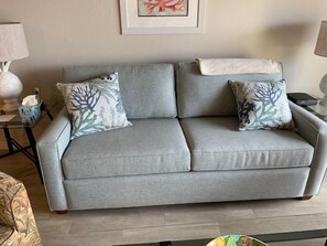 A grey sofa with two patterned throw pillows, a framed picture on the wall, and a small table with a lamp and a tissue box.