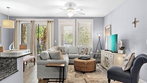 Una sala de estar moderna con un sofá gris, un sillón oscuro, un taburete redondo de mimbre y un televisor de pantalla plana sobre un mueble blanco.