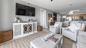 Ein Wohnzimmer mit Flachbildfernseher, weißen Möbeln und einer Küche im Hintergrund.
