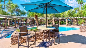 Un espace aménagé au bord de la piscine, avec une table et des chaises sous un parasol bleu, entouré de piliers en pierre et de palmiers.