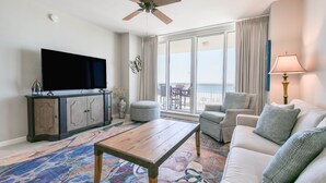 A living room with a flat-screen TV, a ceiling fan, a large window with curtains, a sofa, a coffee table, and a lamp.