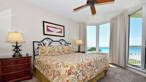 A bedroom with a large bed, two bedside tables, a ceiling fan, a window with curtains, and a framed picture on the wall.