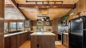 Una cocina con armarios de madera, una isla central, ollas colgantes, un refrigerador negro y vistas a montañas a través de ventanas amplias.
