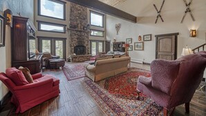 Ein geräumiges Wohnzimmer mit Kamin aus Stein, großen Fenstern und einem roten Teppich mit Muster.