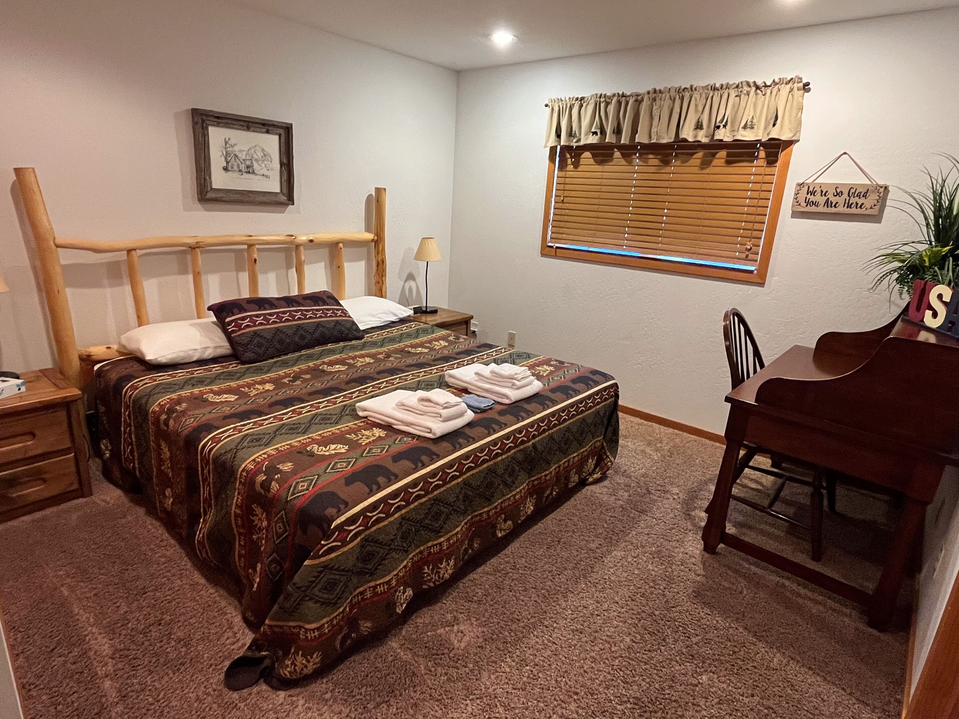 Un dormitorio con cama, escritorio, silla, lámpara, un cuadro enmarcado, ventana con persianas y un cartel en la pared.