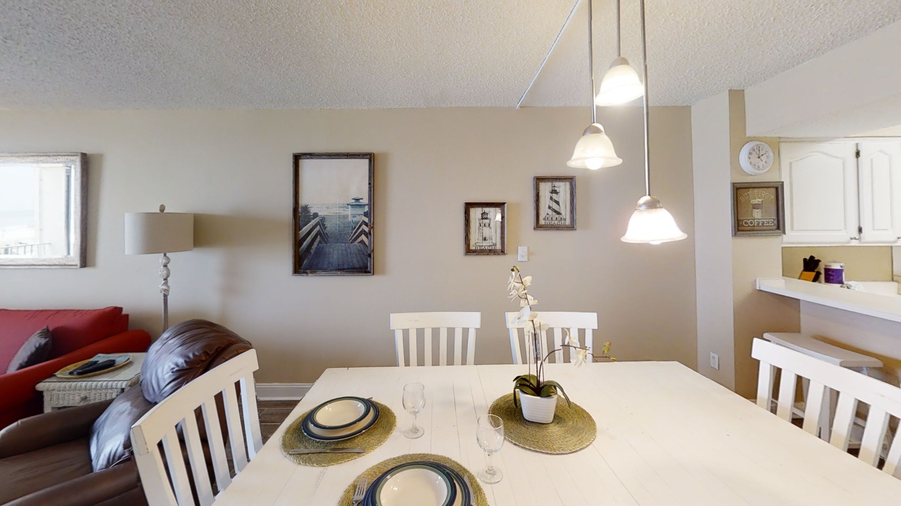 A dining area with a white table set for four, surrounded by white chairs. There are framed pictures on the wall, a lamp, and a coffee station in the background.
