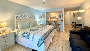 A bedroom with a bed, bedside tables, a ceiling fan, and a kitchenette in the background. - Sugar Beach 122 by ALBVR - Nice Studio Condo with New Sofa, AC and Smart TV (Orange Beach)