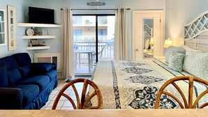 A room with a blue couch, a bed with a headboard, a ceiling fan, a TV, and a view of a balcony with white railings. - Sugar Beach 122 by ALBVR - Nice Studio Condo with New Sofa, AC and Smart TV (Orange Beach)