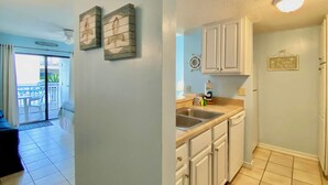 A kitchen with white cabinets, a sink, and a countertop. There is a clock on the wall and a sign that says "Life is better at the beach." - Sugar Beach 122 by ALBVR - Nice Studio Condo with New Sofa, AC and Smart TV (Orange Beach)