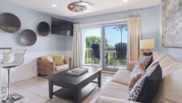 A spacious living room with a sofa, a coffee table, and a wicker chair. There is a large window with a view of the outdoors. - Breakers 110 - 1st Floor Ocean Front Condo (Hilton Head Island)