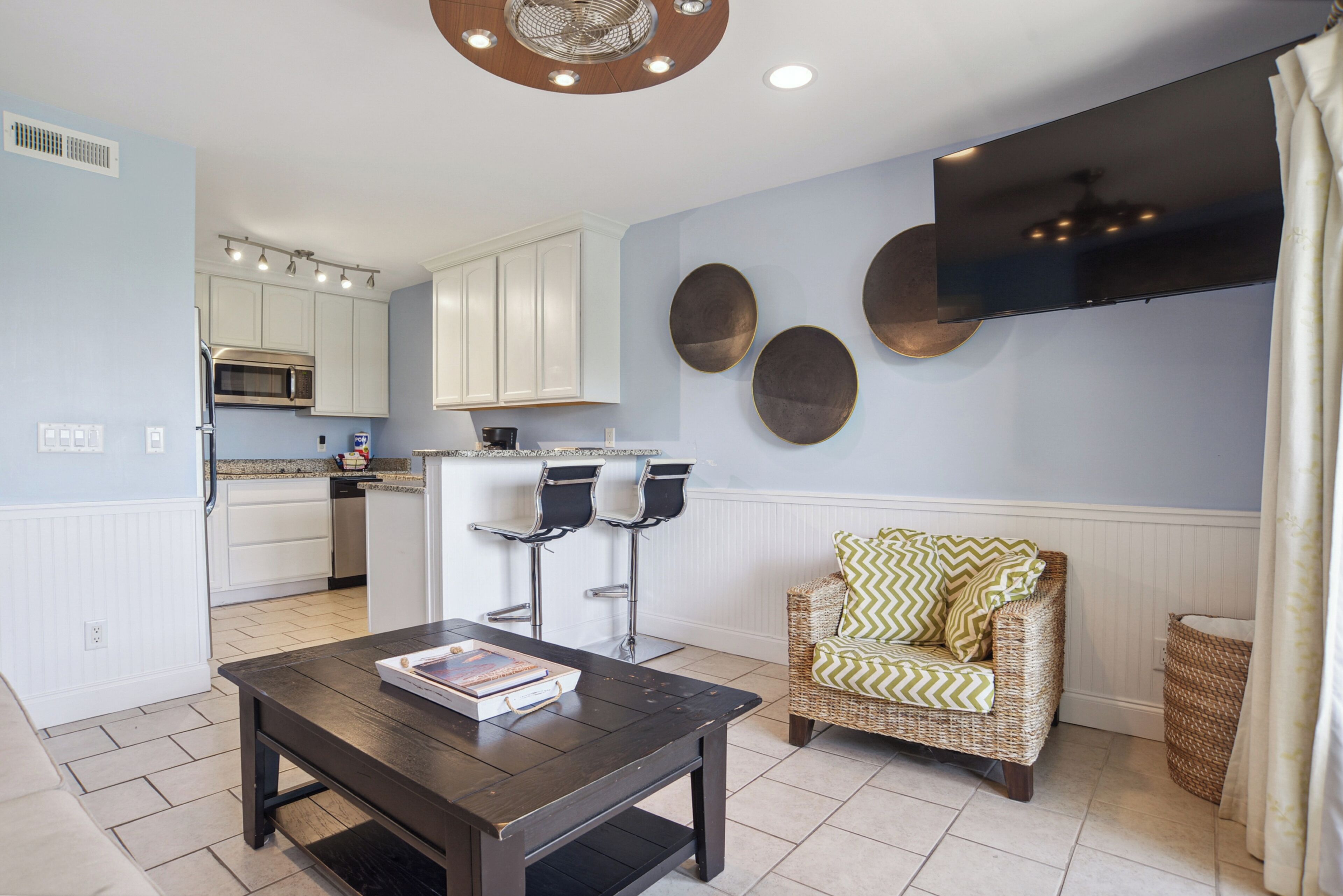 A modern kitchen with white cabinets, a microwave, and a coffee machine. There is a dining area with a dark wooden table and a wicker chair.