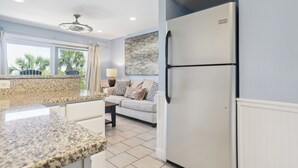 A modern kitchen with granite countertops, a stainless steel refrigerator, a dining area with a white sofa, and a large window with a view of palm trees. - Breakers 110 - 1st Floor Ocean Front Condo (Hilton Head Island)