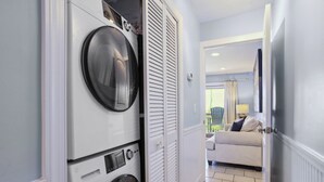 A laundry room with stacked washers and dryers, a white door, and a view into a living area with a couch and a lamp. - Breakers 110 - 1st Floor Ocean Front Condo (Hilton Head Island)