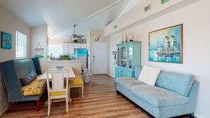 A living room with a sofa, a dining table with chairs, a kitchen area, and a hallway leading to a bedroom. - Pet Friendly, Boardwalk, Glimpse of Ocean from Deck (Port Aransas)