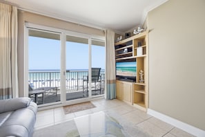 A living room with a sofa, a flat-screen TV, a balcony with a view of the beach, and a wooden entertainment unit.