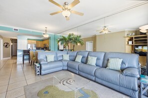 A living room with a sectional sofa, ceiling fans, a kitchen in the background, and a large potted plant.