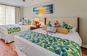 Una habitación de hotel con dos camas, cada una con una colcha estampada con motivos tropicales, una mesita de noche con lámpara y un cuadro con un atardecer sobre el océano en la pared.