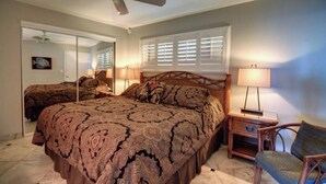 Ein Schlafzimmer mit einem großen Bett, Deckenventilator, Nachttisch mit Lampe, einem Sessel und einem Fenster mit Jalousien.