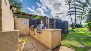 Ein Außenbereich mit Grillplatz, einer Steintheke, zwei Grills und einem von einem Zaun umgebenen Garten mit Palmen.