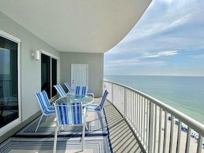 Een balkon met een glazen tafel en blauw gestreepte stoelen, met uitzicht op een strand.