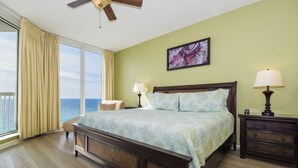 Un dormitorio con una cama grande, una mesita de noche, un ventilador de techo, una ventana con cortinas y vista al océano.