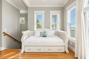A bedroom with a white bed, two windows with blinds, a wooden staircase, and a view of greenery outside.