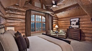 Ein Schlafzimmer in einer Blockhütte mit einem großen Bett, einem Fenster mit Vorhängen, einem Sessel, einer Lampe und einer Kommode.