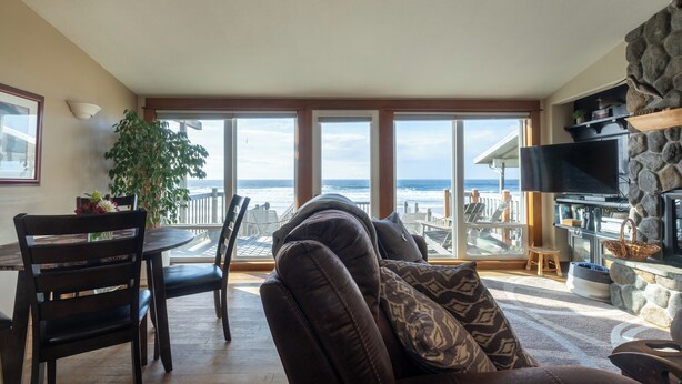 A cozy living room with a large window overlooking the ocean, a stone fireplace, and a dining area with chairs and a table.