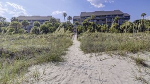 Un percorso che conduce a un hotel fronte spiaggia con diversi piani e balconi, immerso tra palme e vegetazione costiera.