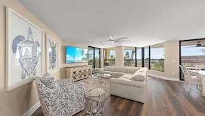 Un salón espacioso con un sofá grande, una mesa de centro y un televisor. Amplias ventanas ofrecen vistas al exterior.