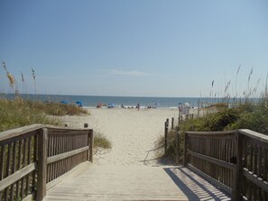 Walk along the boardwalk to the sandy beach, flanked by railings and surrounded by lush vegetation.