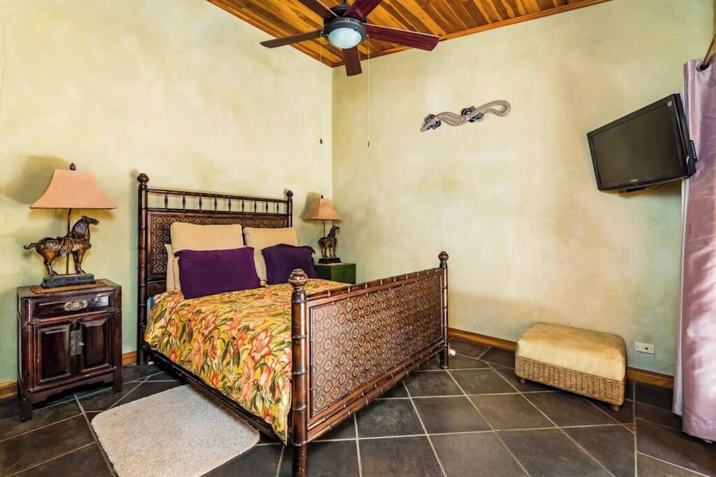 Quarto com cama de madeira, duas mesas de cabeceira, um criado-mudo, um banquinho de palha, um ventilador de teto e uma TV fixada na parede.