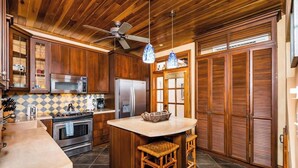 A kitchen with wooden cabinets, a stainless steel refrigerator, a microwave, and a stove. There is a ceiling fan and a hanging light fixture. - Villa Buho✰Rooftop Jacuzzi & Private Pool ✰Across from Beach (Tamarindo)