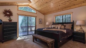 A bedroom with a large bed, a bench, a dresser, and a nightstand. There are two windows providing natural light and a view of trees.