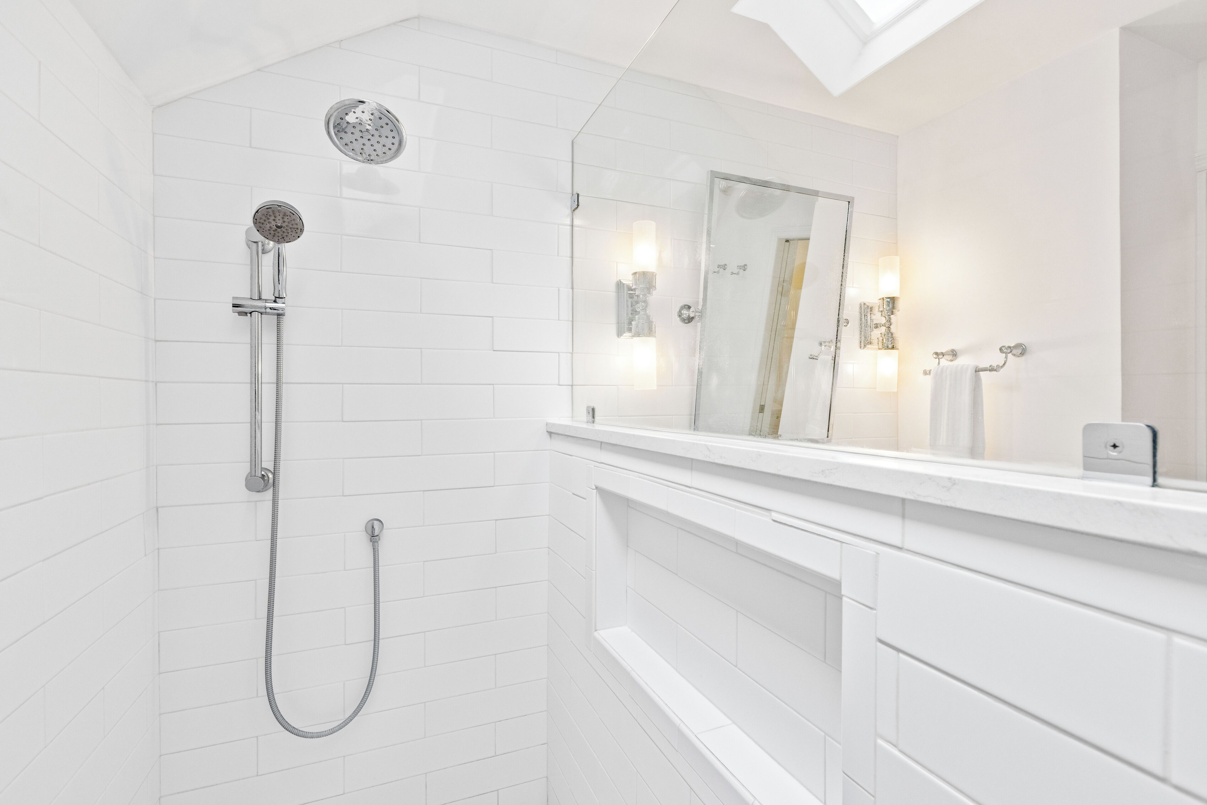 A modern bathroom with white tiled walls, a glass shower enclosure, a white vanity with a mirror, and a towel rack.