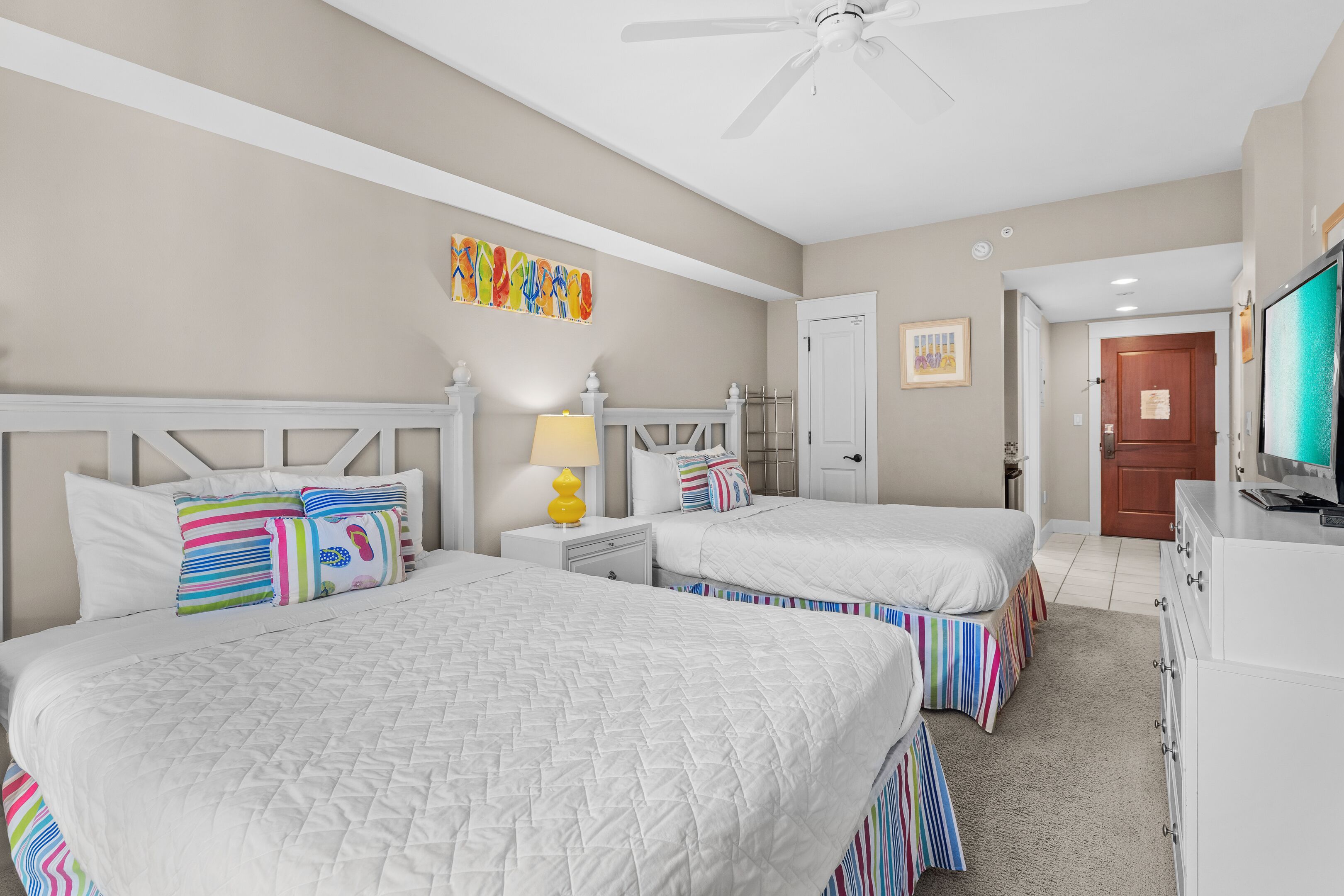 A hotel room with two beds, a ceiling fan, a TV, and a door leading to another room.