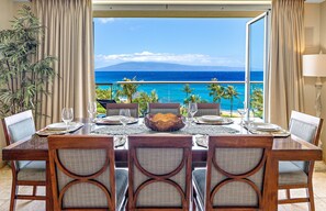 A dining table with chairs, a bowl, and wine glasses, set against a backdrop of a beach and ocean view.
