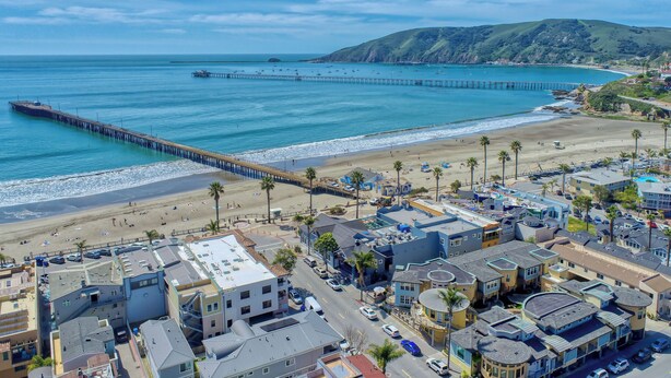 Un pueblo costero con un muelle que se adentra en el océano, propiedades frente a la playa y un paisaje montañoso al fondo.