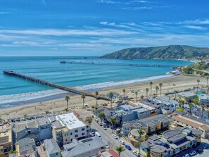 Un pueblo costero con un muelle que se adentra en el océano, propiedades frente a la playa y un paisaje montañoso al fondo.